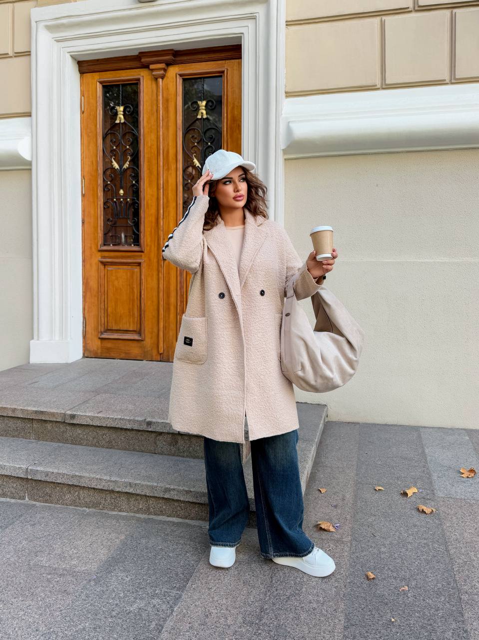 Woman in a beige coat holding a coffee cup and a bag, standing in front of a building entrance.