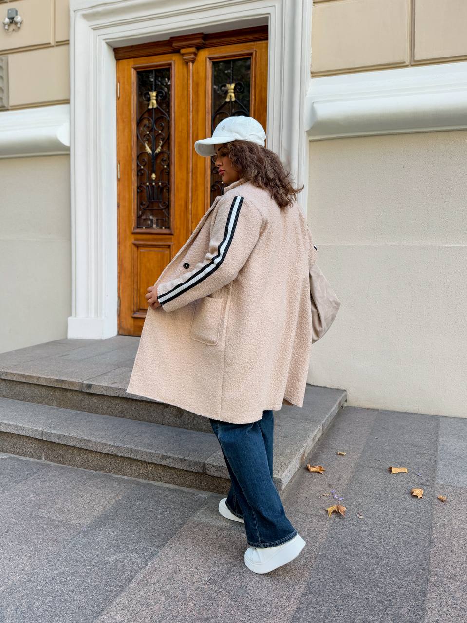 Person wearing a beige coat with black and white stripes, blue jeans, and white sneakers standing in front of a building entrance.