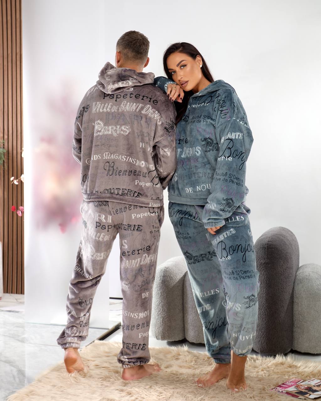 Two people wearing matching pajama sets with text design in a living room.