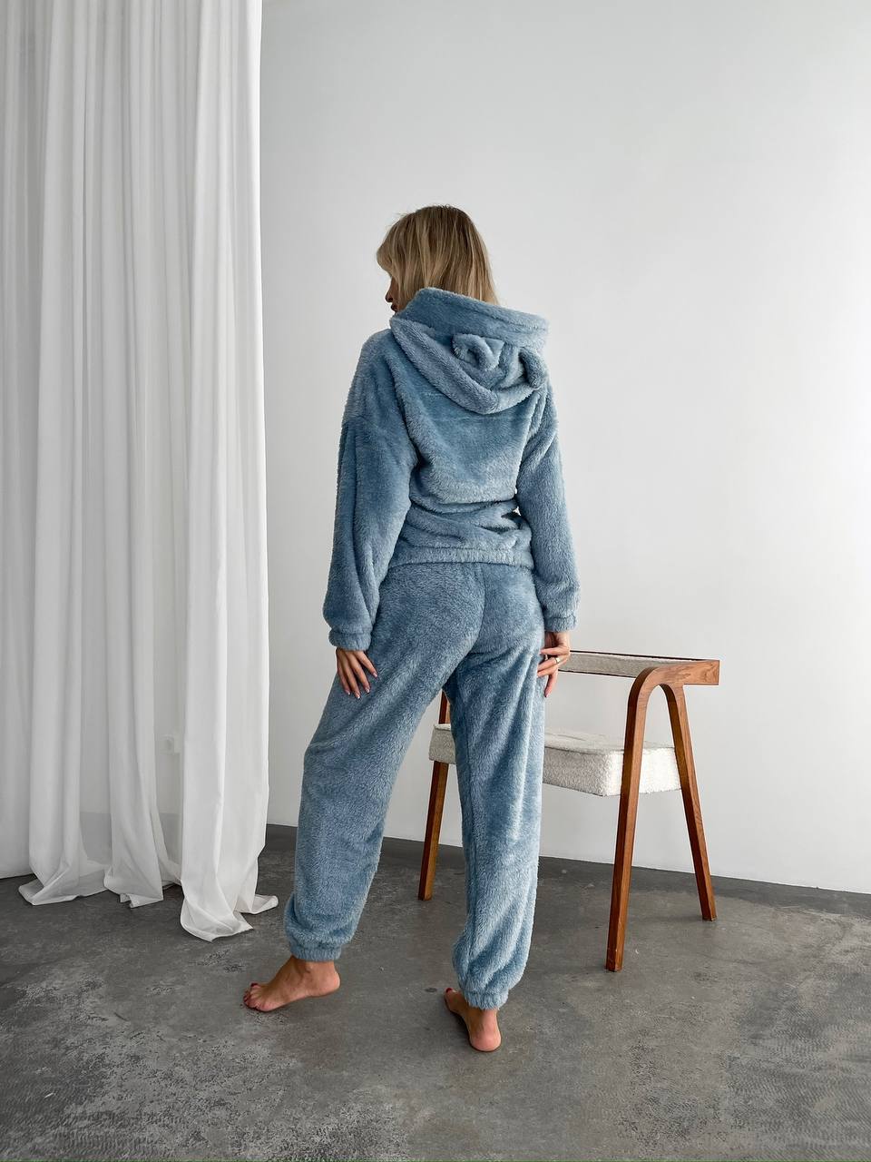 Person wearing a blue tracksuit standing in a minimalistic room with white curtains and a wooden stool.