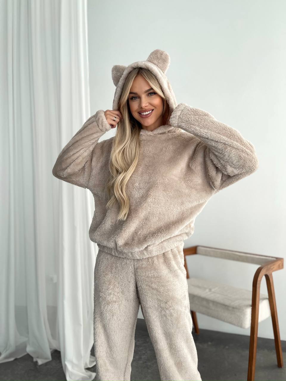 Woman wearing a beige fleece hoodie and pants with bear ears in a minimalistic room.