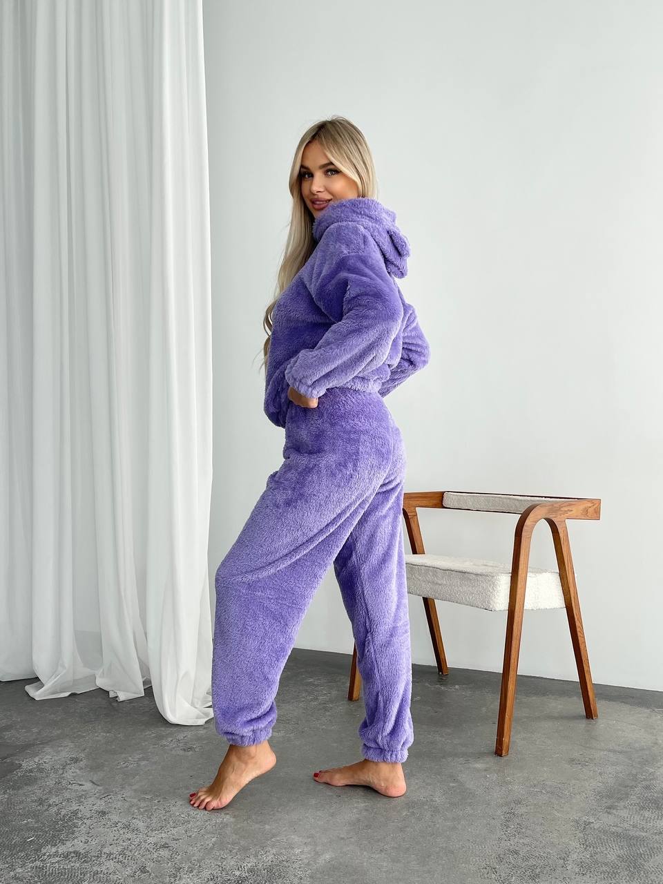 Woman wearing a purple fleece onesie standing in a room with a white curtain and wooden chair.