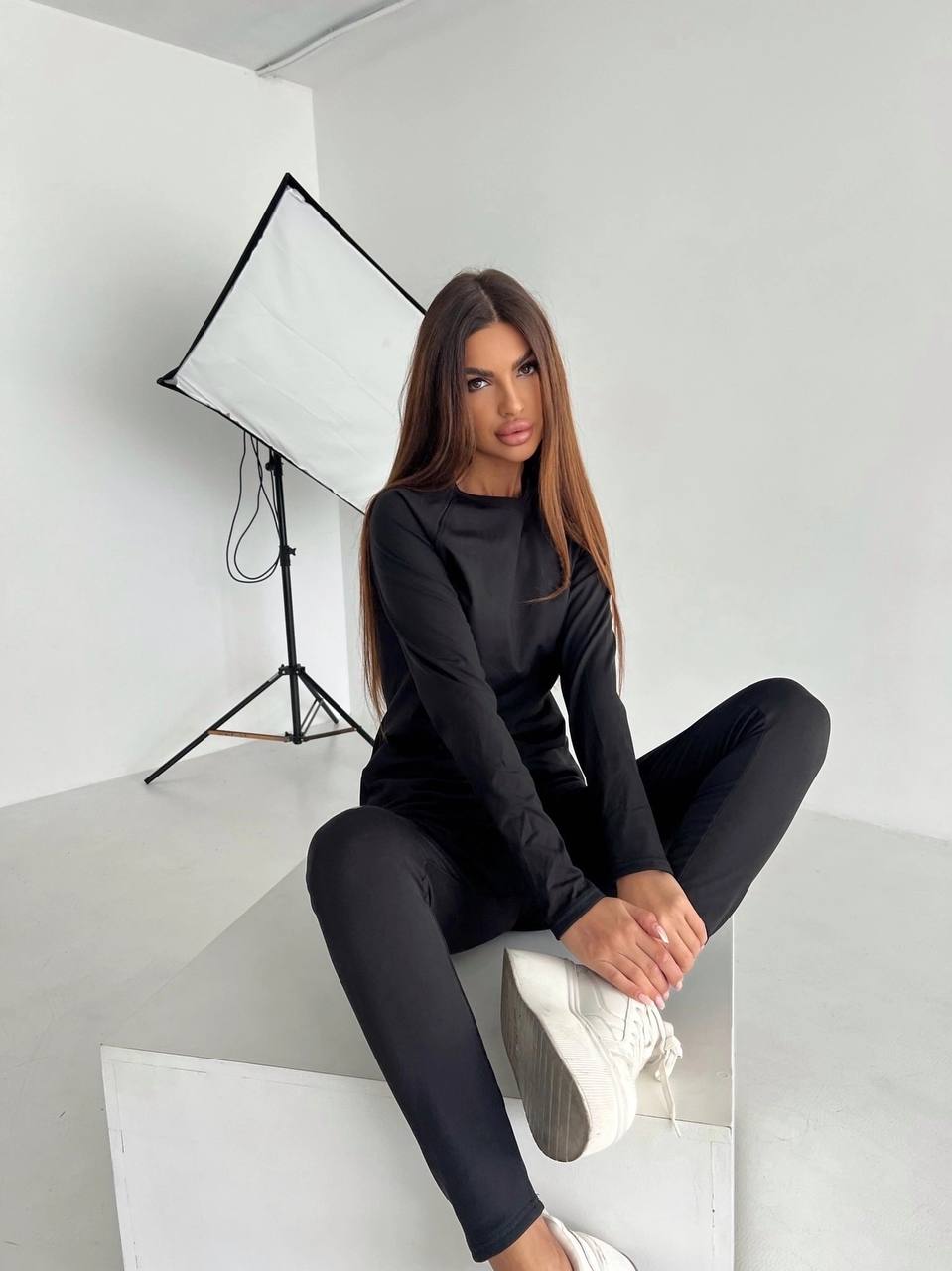 Woman in black outfit sitting on a white surface with studio lighting equipment in the background.