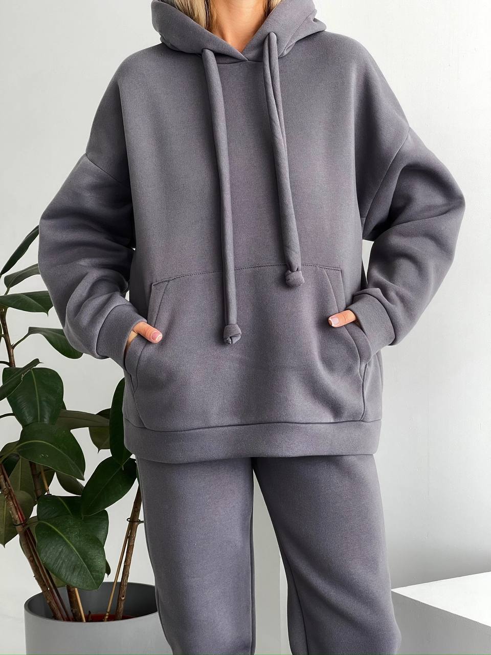 Person wearing a gray hoodie and pants with a plant in the background