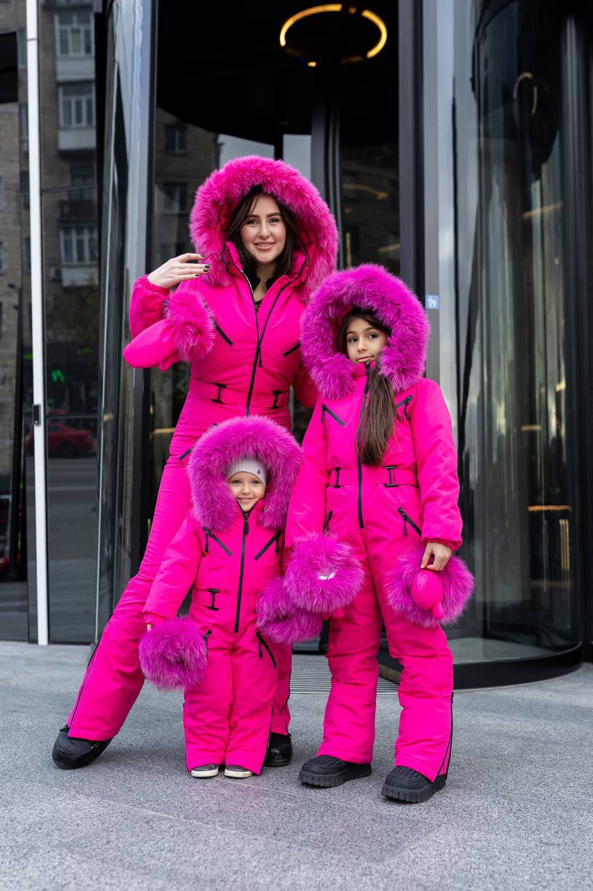 Three people in matching bright pink snow suits with fur hoods standing in front of a glass door.