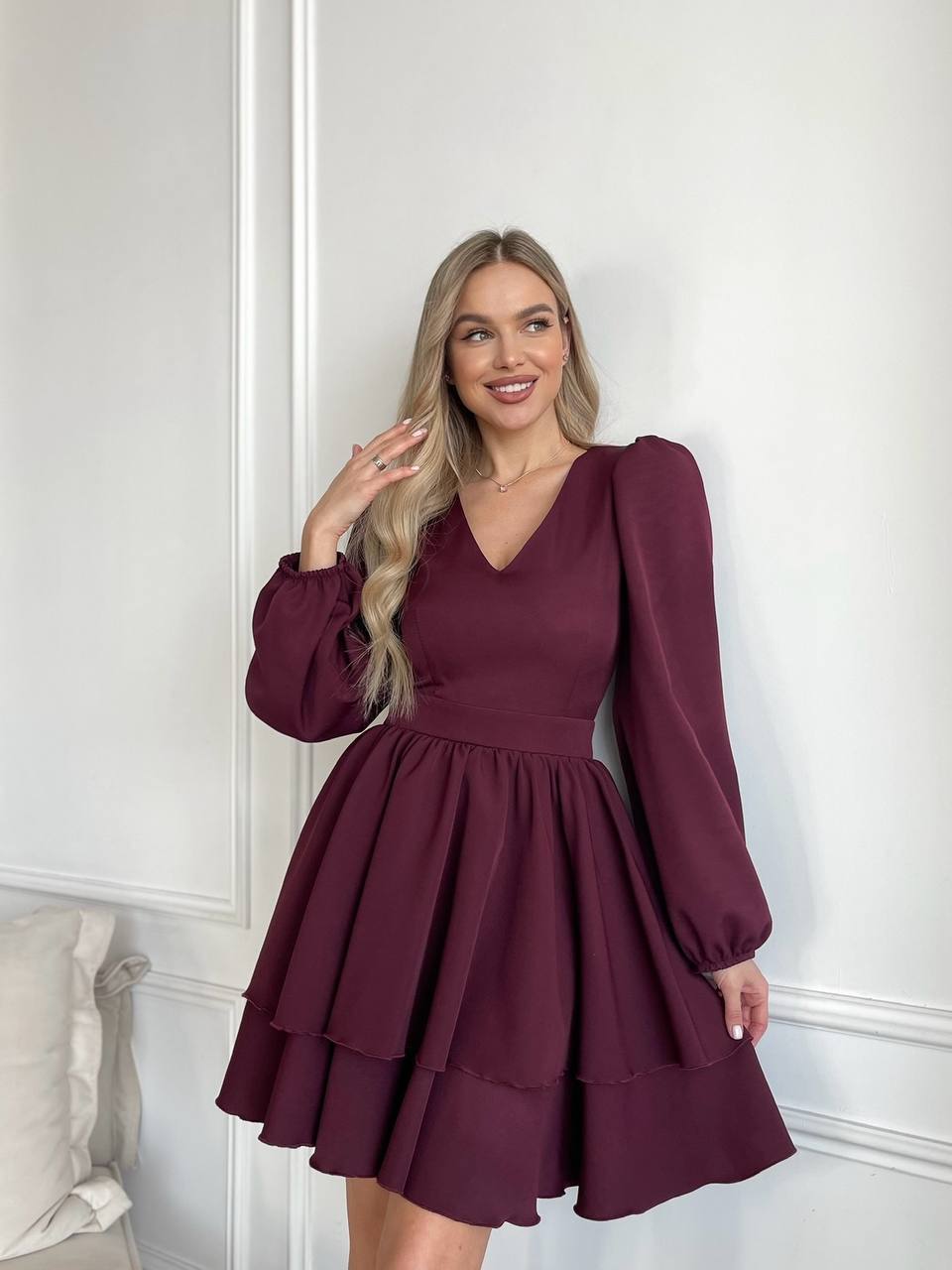 Woman wearing a burgundy dress with long sleeves against a white wall.