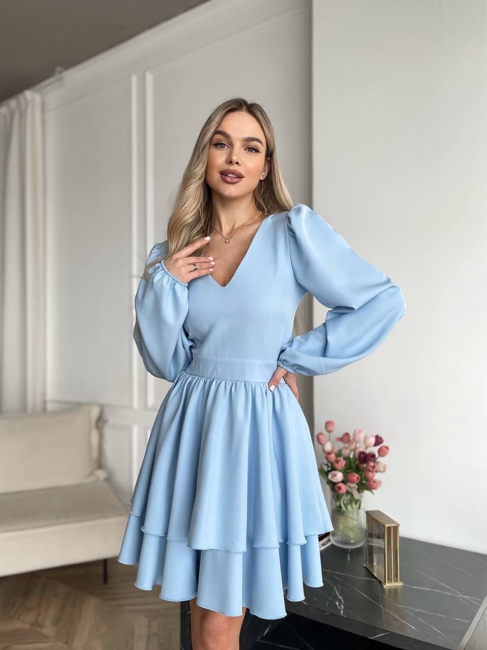 Woman wearing a light blue dress standing in a room with a couch and flowers.