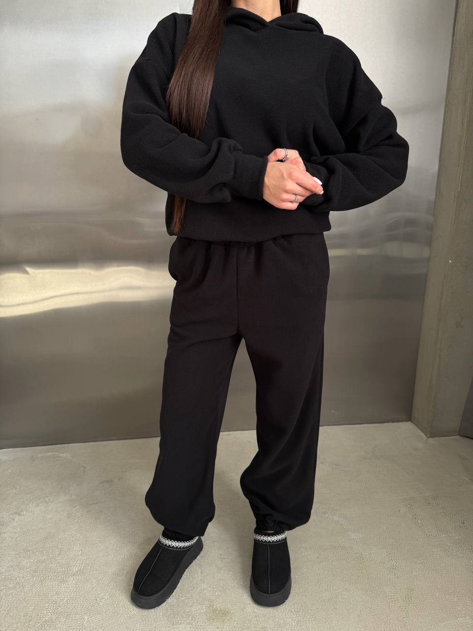 Person wearing a black hoodie and pants with black shoes in a neutral setting
