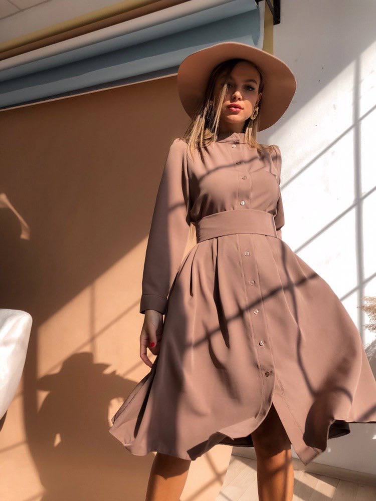 Woman wearing a long beige coat and hat standing against a wall with sunlight casting shadows.