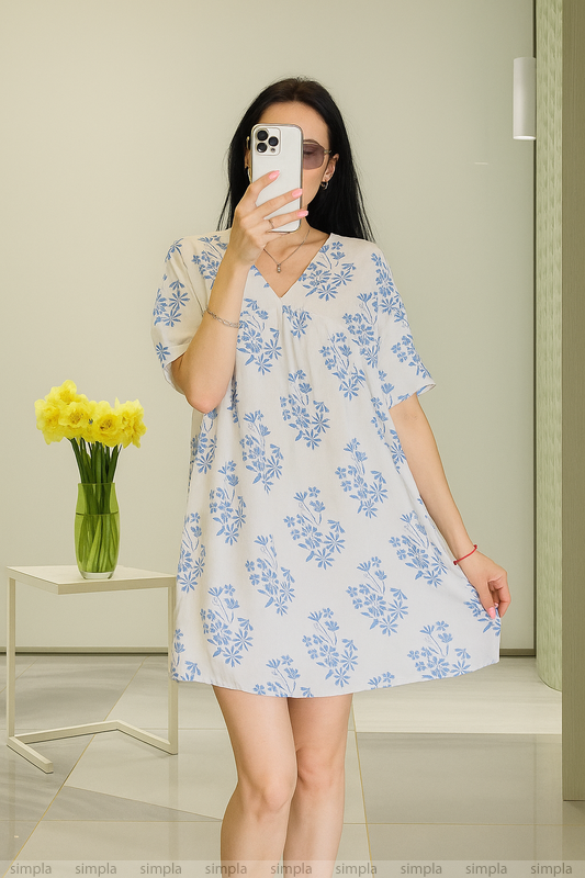 Woman wearing a floral dress taking a mirror selfie in a room with a vase of flowers on a table.