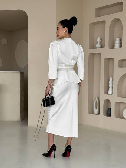 Woman in a white outfit holding a black handbag in a minimalistic room.