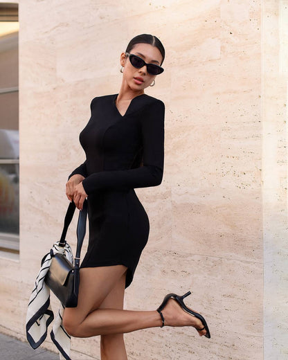 Woman in a black dress holding a handbag against a beige wall.