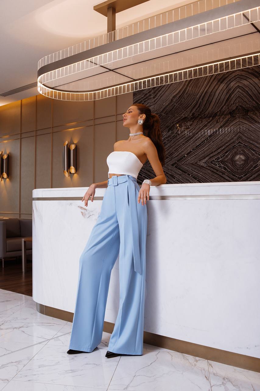 Woman in a white top and light blue pants standing in an elegant interior setting.
