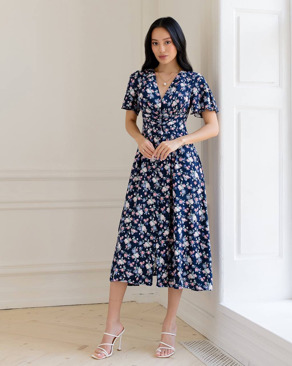 Woman wearing a floral dress standing against a light-colored wall.