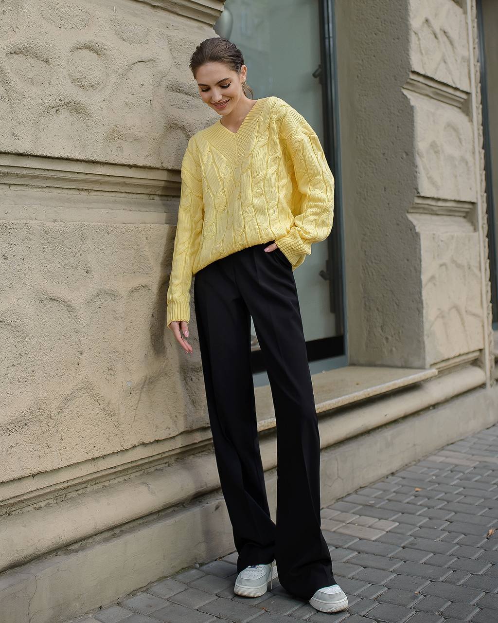 Woman wearing a yellow sweater and black pants standing against a stone wall.