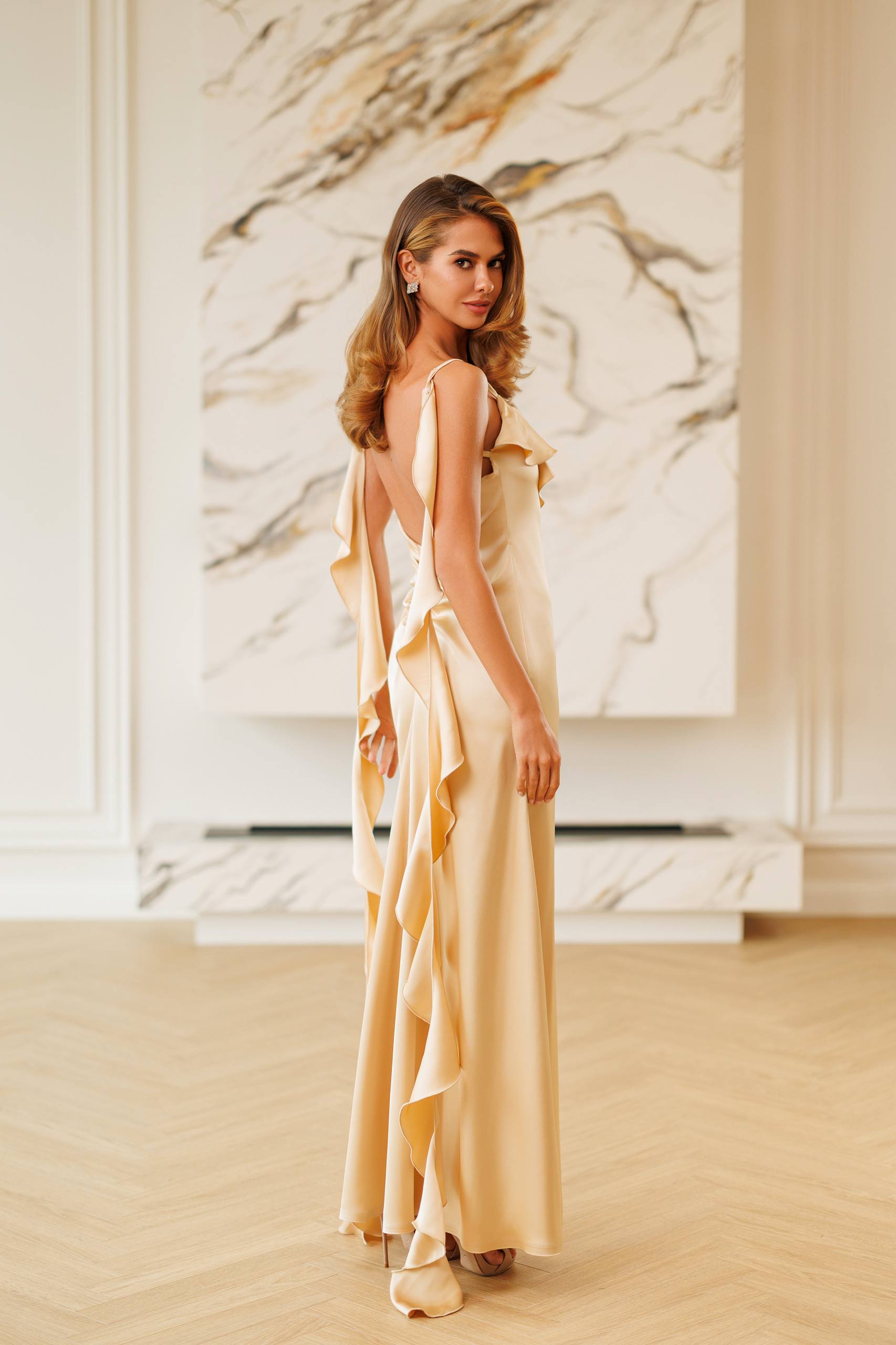A woman modeling a floor-length cream satin gown with dramatic draped ruffle details, photographed in a bright, elegant room with a marble wall art piece and light wood flooring.