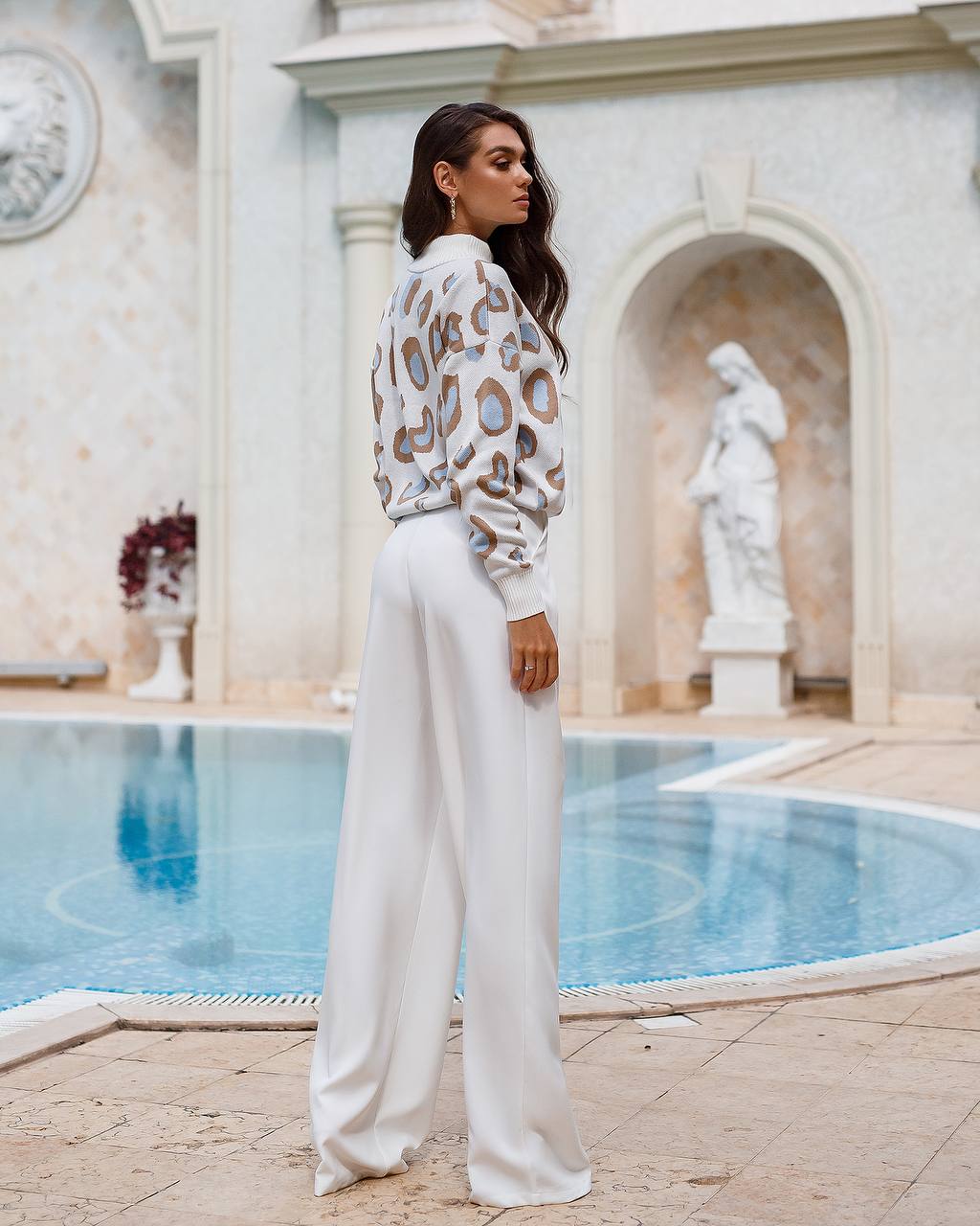 Woman in a leopard print top and white pants standing by a pool with classical architecture.