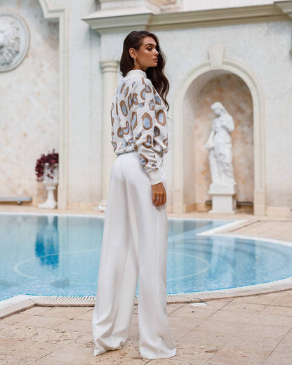 Woman in a leopard print top and white pants standing by a pool with classical architecture.