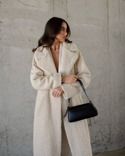 Woman wearing a long cream coat holding a black handbag against a plain background
