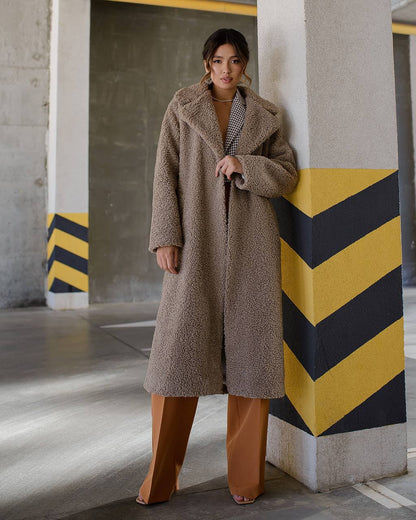 Woman wearing a long brown coat in an urban setting with concrete pillars and yellow and black stripes.