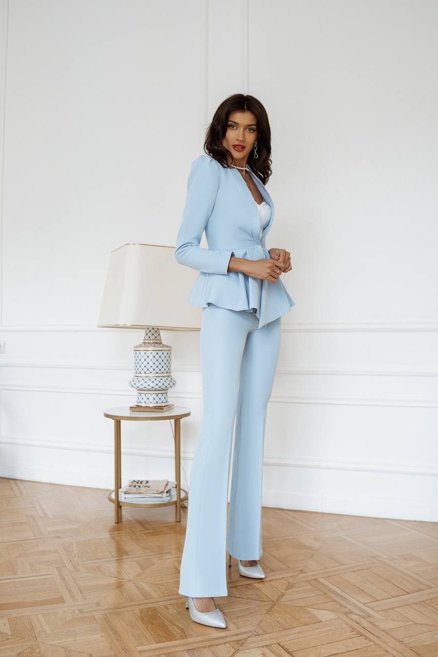 Woman in a light blue suit standing in a room with a lamp and side table.
