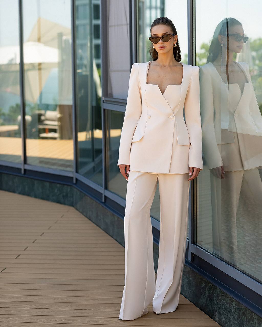 A woman models a light cream double-breasted blazer and matching wide-leg trousers set, styled with sunglasses, standing on a wooden deck next to a glass-walled building.