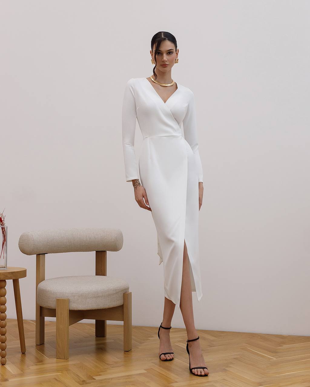 Woman in a white dress standing in a room with a chair and table.