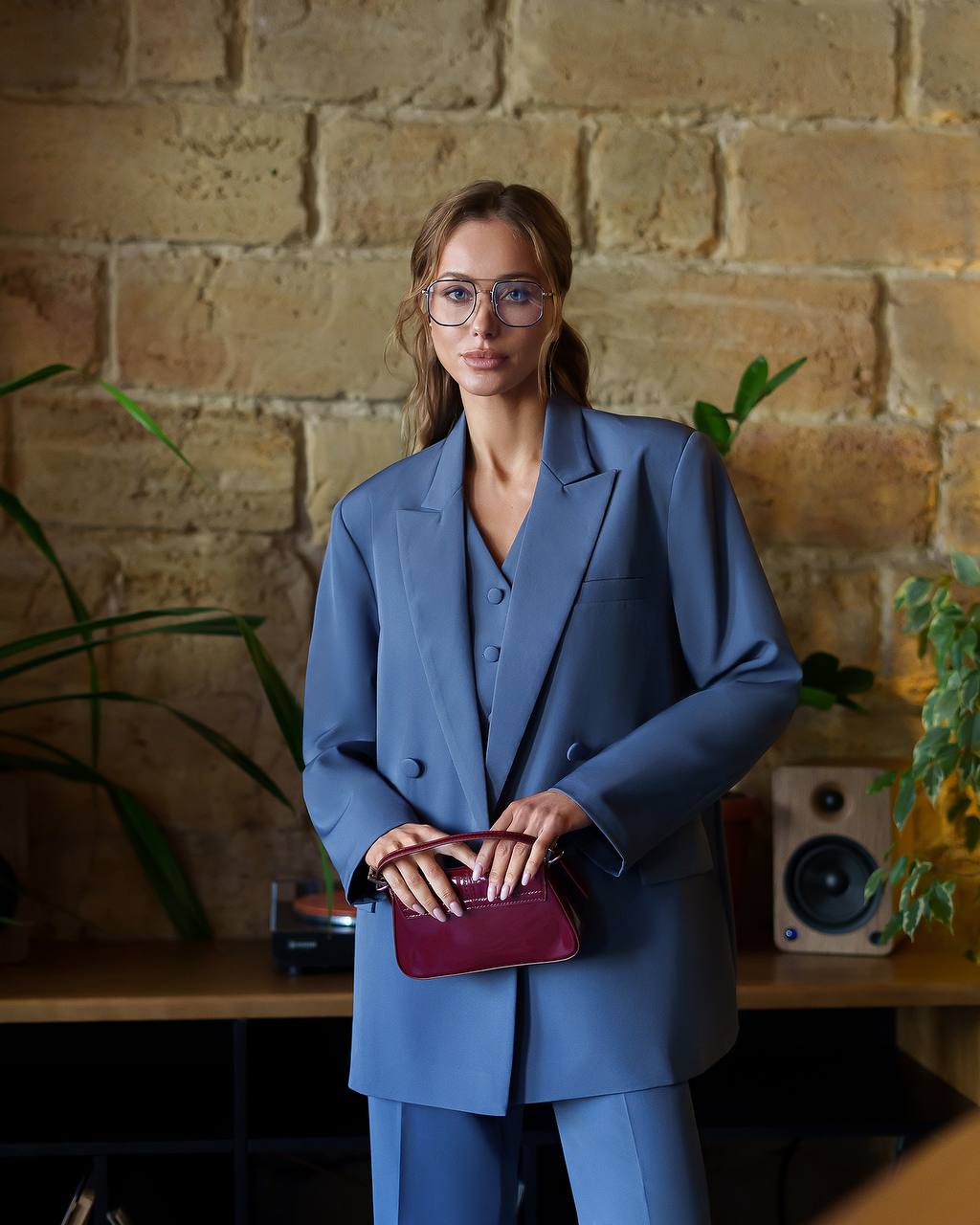 Woman in a blue suit holding a purple clutch against a brick wall.