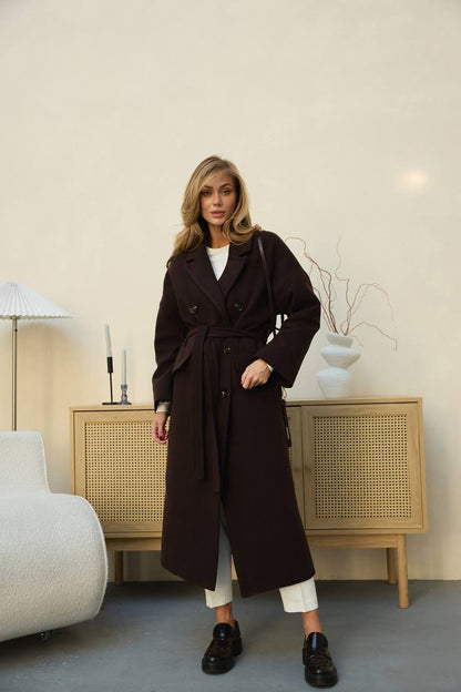 Woman wearing a long brown coat in a room with a light-colored wall and wooden cabinet.