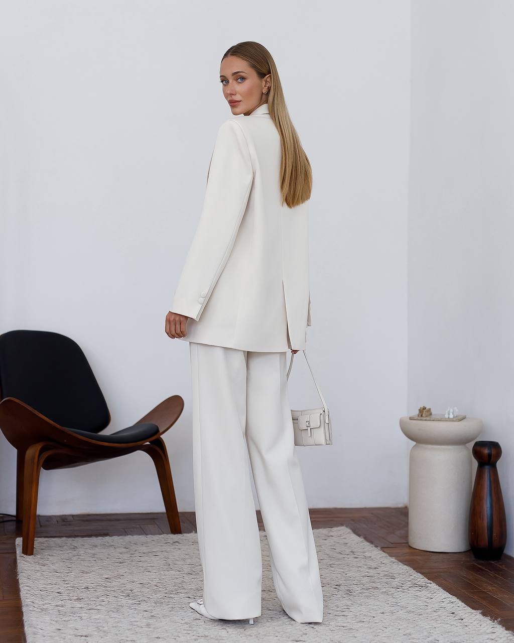 Woman in a white suit standing in a minimalistic room with a chair and table.