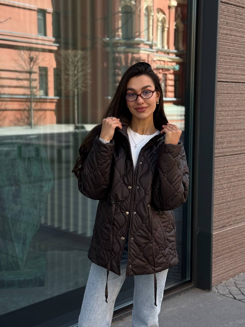 Woman wearing a brown quilted jacket and glasses standing in front of a building.