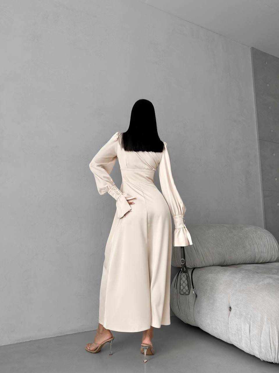 Person wearing a long beige dress standing in a minimalistic room with gray walls and furniture.