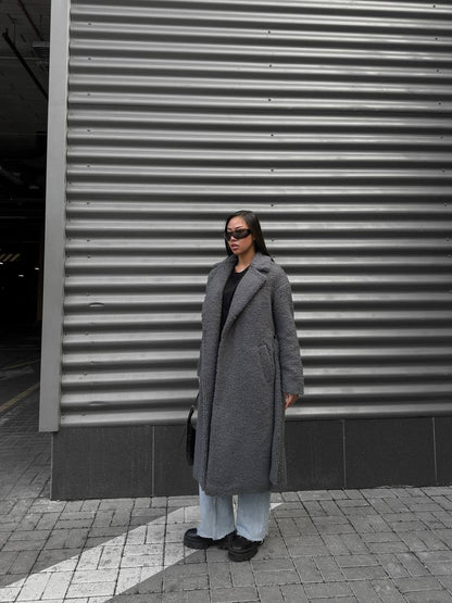 Person wearing a long gray coat standing in front of a corrugated metal wall.