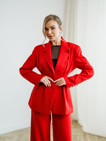 A woman modeling a bright red tailored pantsuit with a structured blazer and matching trousers, worn over a black top, in a bright indoor setting with white walls and light wood flooring.