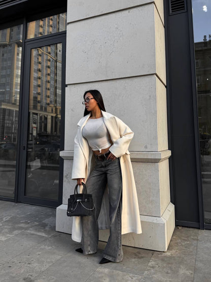 Woman in a white coat and gray pants standing against a building.