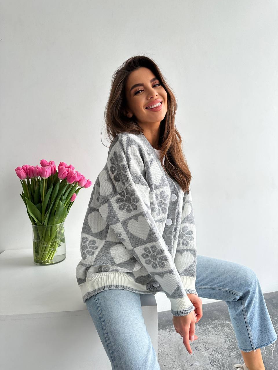 Woman wearing a patterned sweater sitting next to a vase of pink tulips on a white surface.
