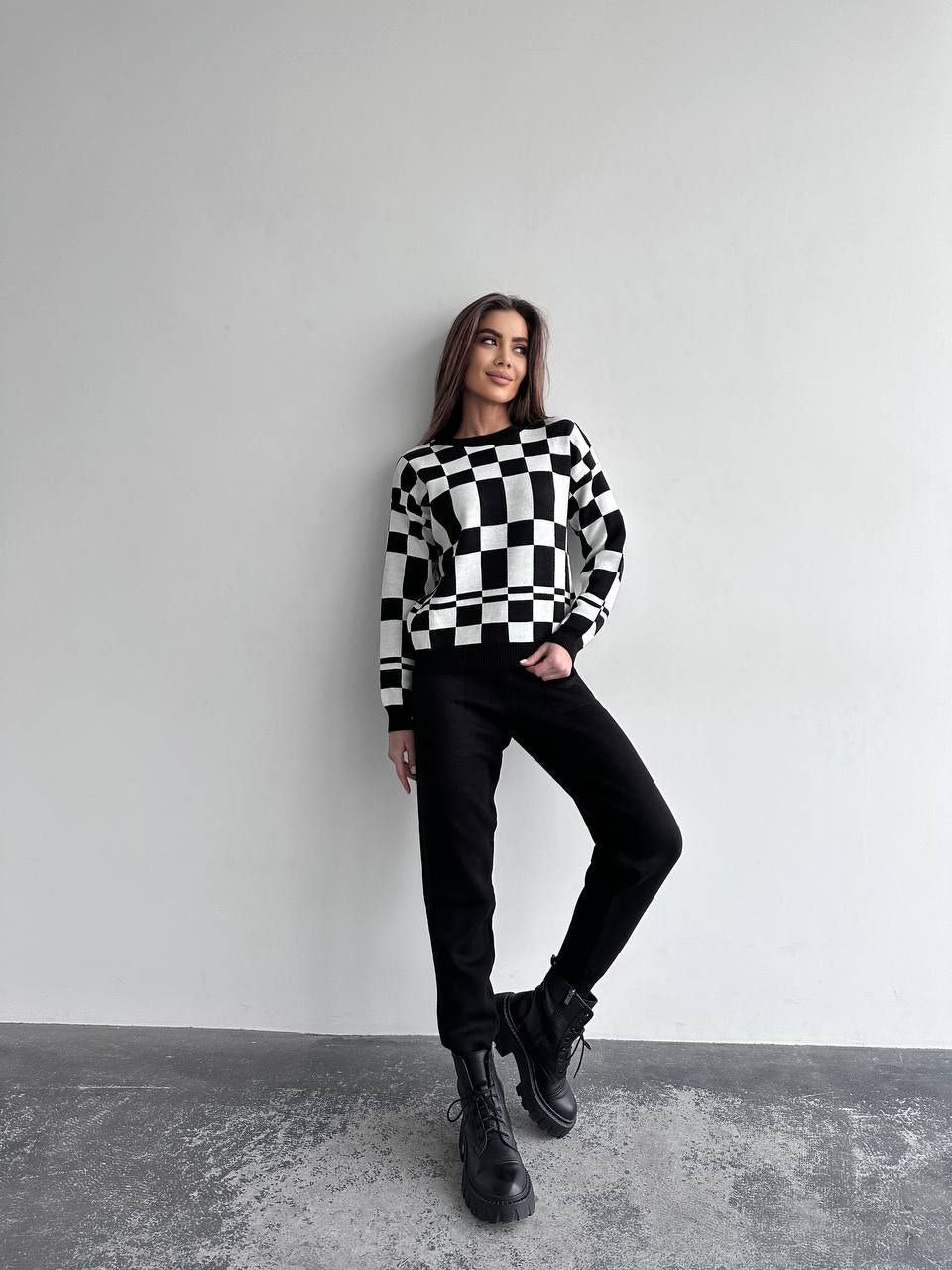 Woman wearing a black and white checkered sweater with black pants and boots against a gray background