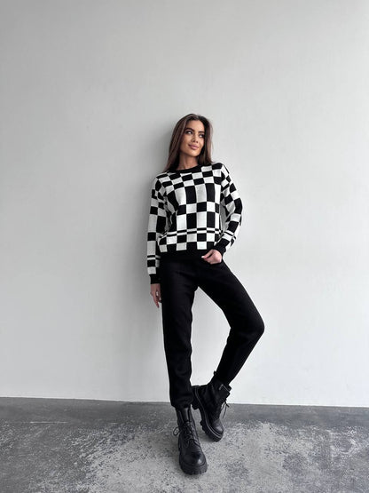 Woman wearing a black and white checkered sweater with black pants and boots against a gray background