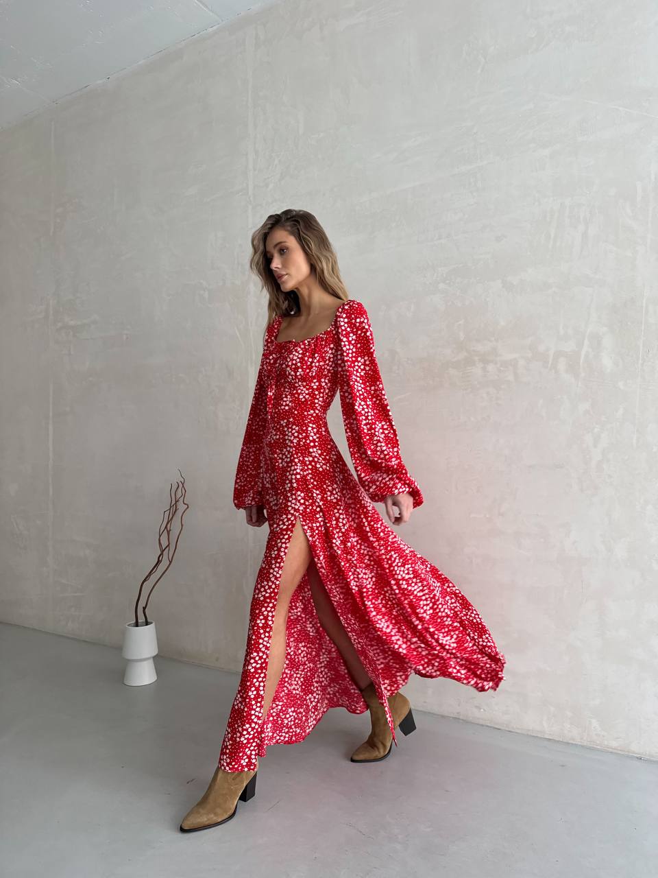 Woman wearing a red floral dress with a slit, standing against a plain white wall.