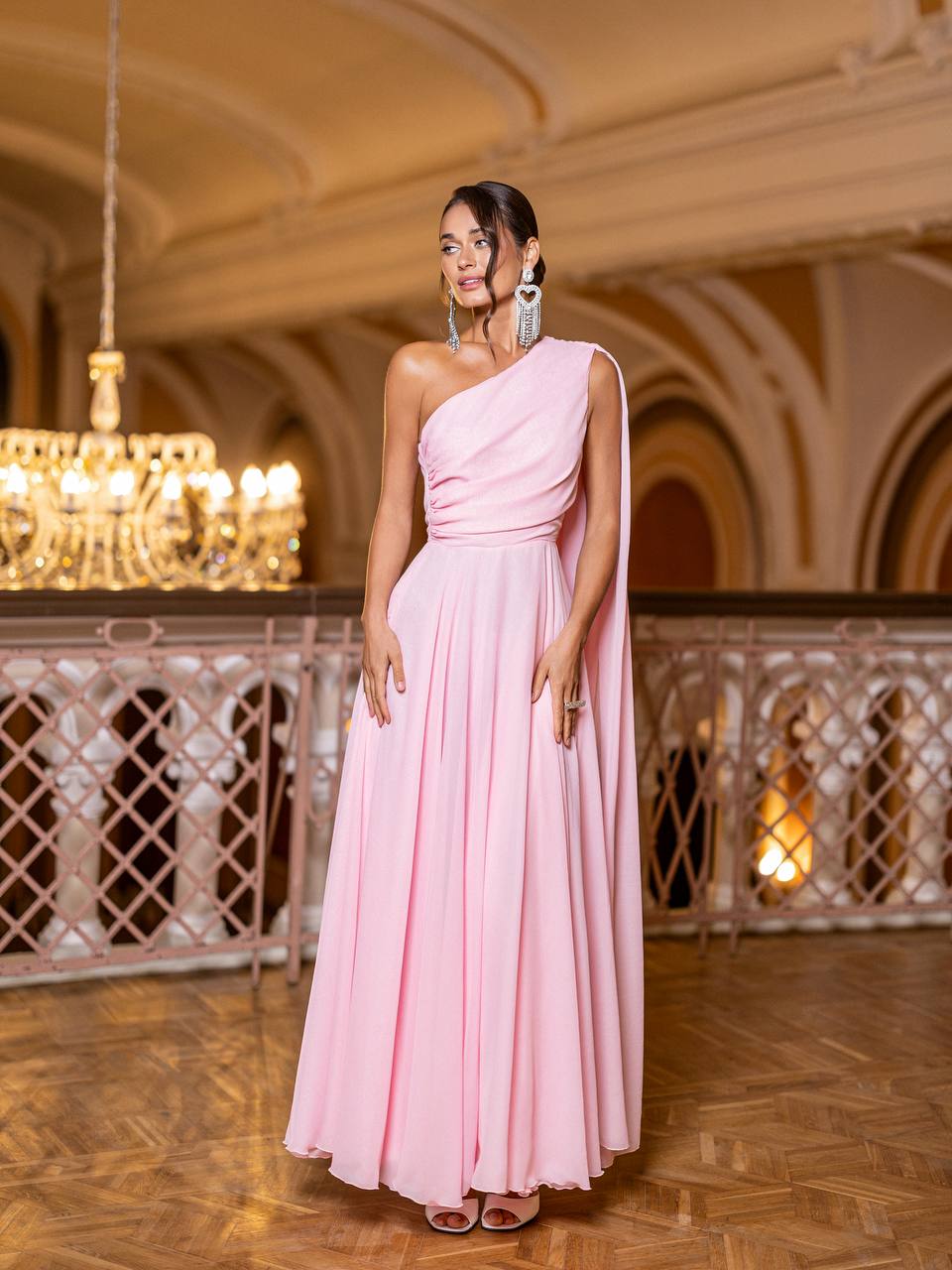 Woman in a pink one-shoulder dress standing in an elegant indoor setting with chandeliers.