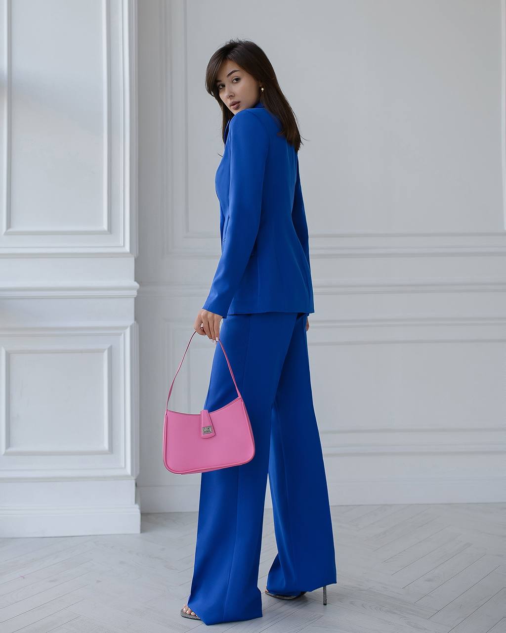 Woman in a blue outfit holding a pink handbag against a white paneled wall.