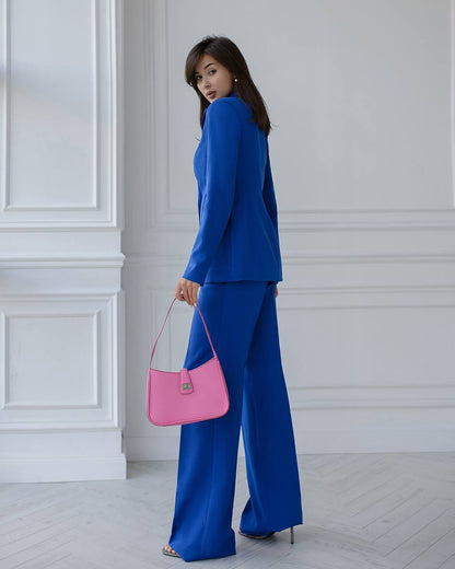 Woman in a blue outfit holding a pink handbag against a white paneled wall.