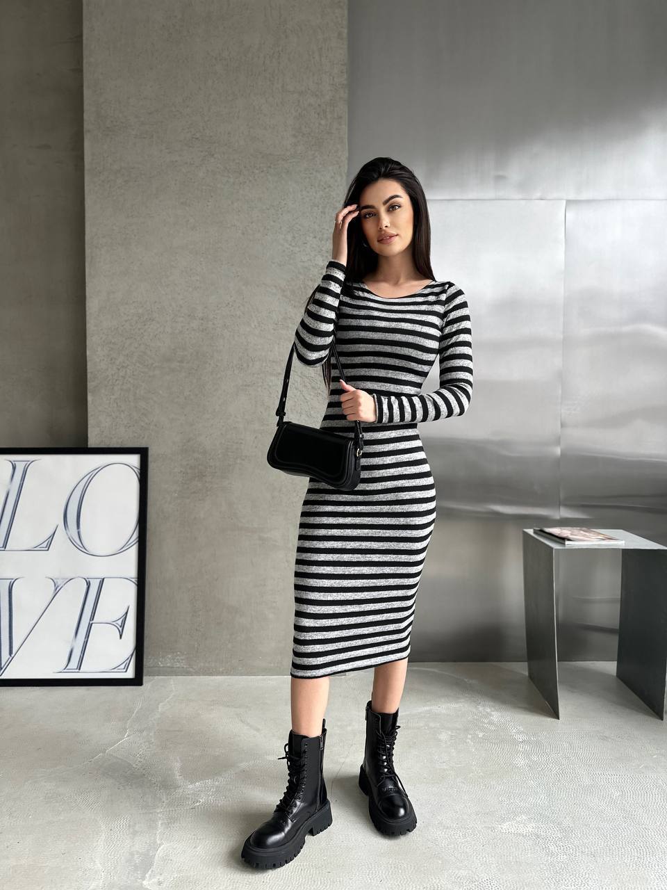 Woman in a black and white striped dress with black boots standing against a gray wall.
