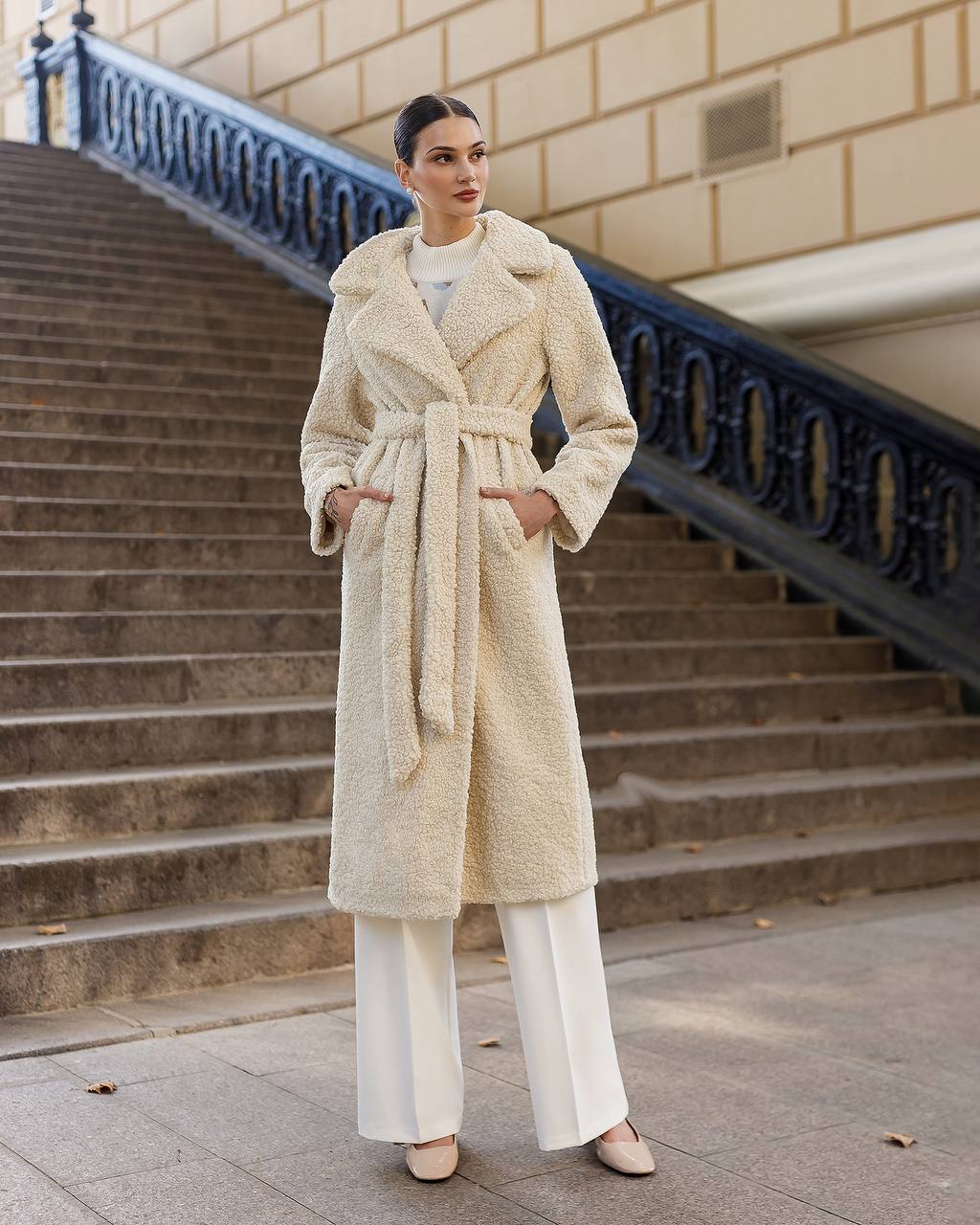 Woman wearing a cream coat standing on stone steps with a building in the background