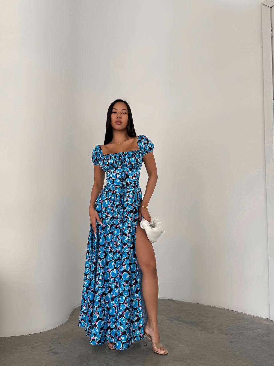 Woman in a blue floral dress with a white clutch against a plain background