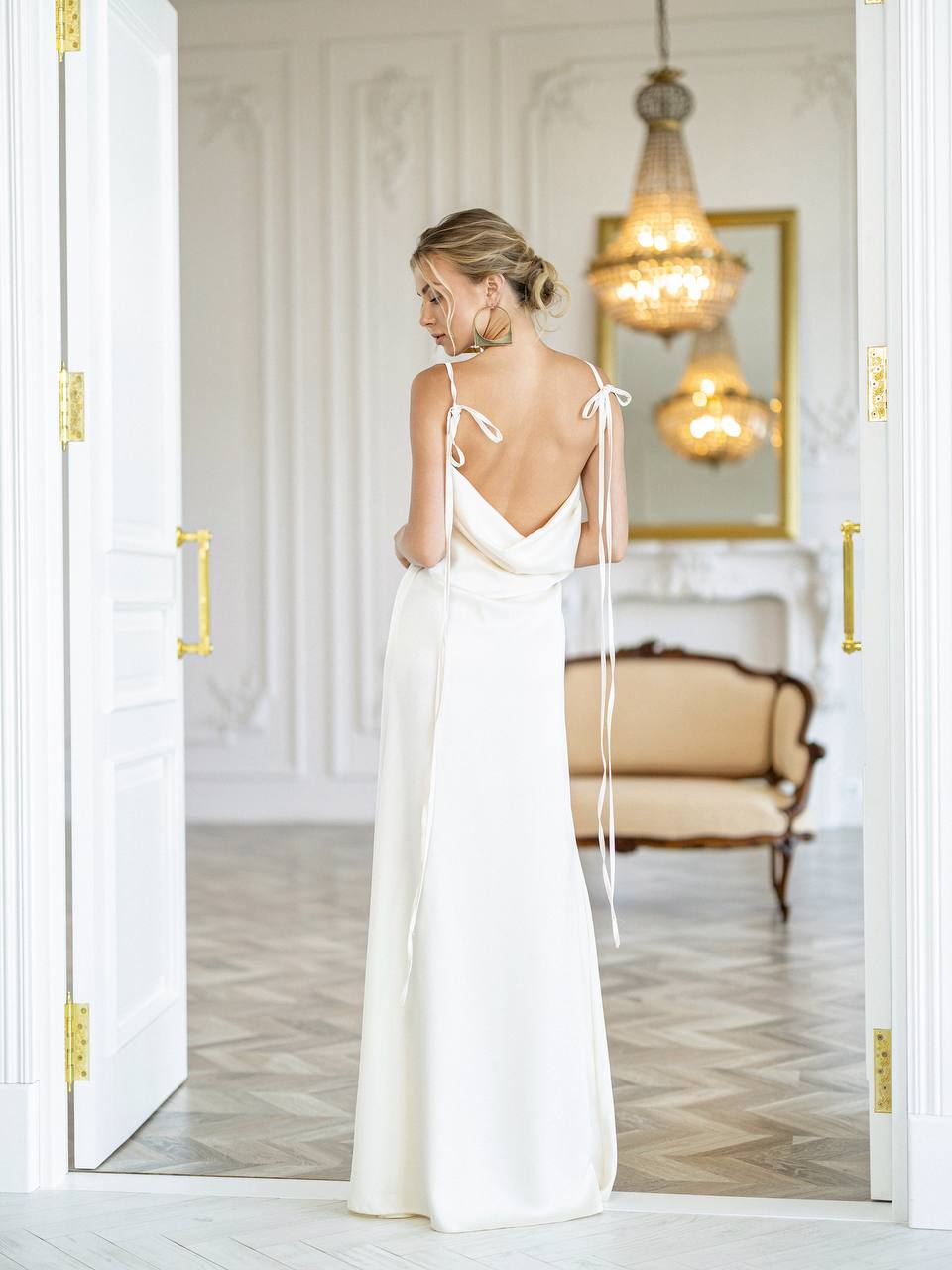 A woman stands with her back to the camera wearing a floor-length white bridal gown with thin tie-back straps, photographed in a bright, elegant room with white walls, gold hardware, and a chandelier reflection.