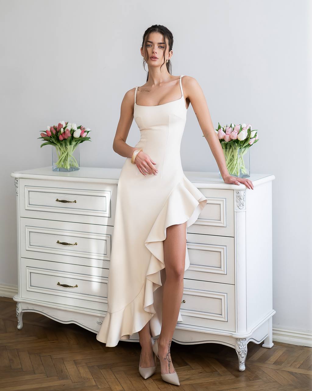 Woman in a white dress standing next to a white dresser with floral arrangements.