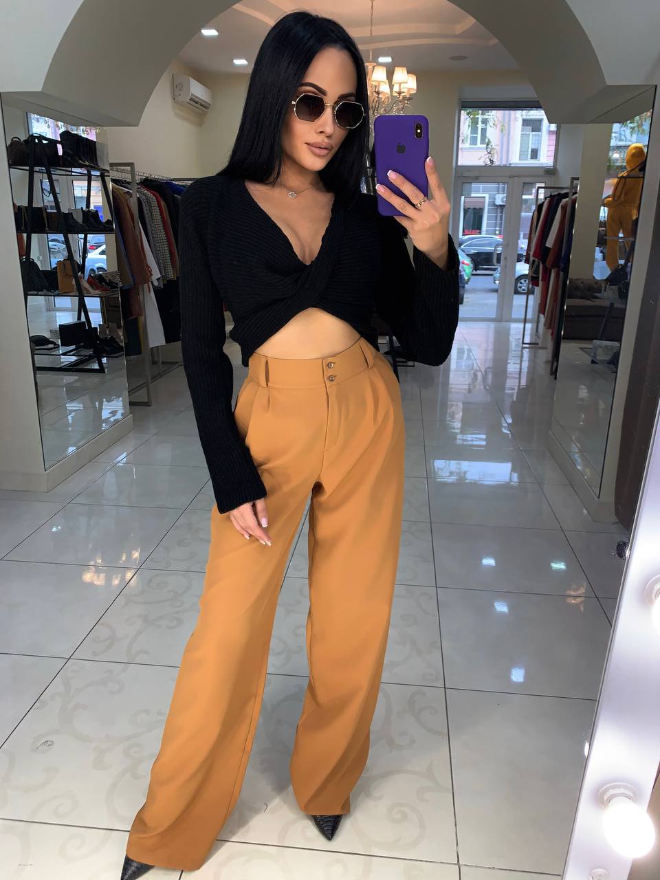 Woman taking a mirror selfie wearing a black top and mustard yellow pants in a store.