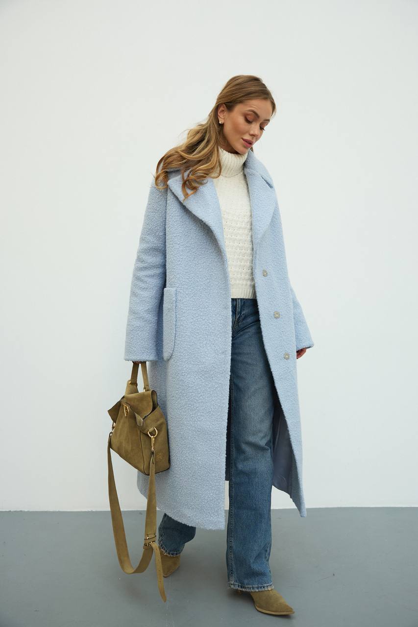 Woman wearing a light blue coat, white sweater, and jeans, holding a brown bag against a white background.