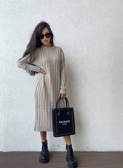 Woman wearing a long-sleeve ribbed dress holding a Balmain bag against a white wall.